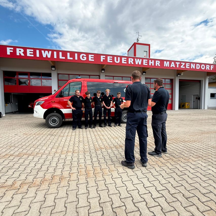 Eine Gruppe von Feuerwehrleuten steht vor einer Feuerwache, mit einem roten Van hinter ihnen. Das Gebäude heißt Freiwillige Feuerwehr Matzendorf.