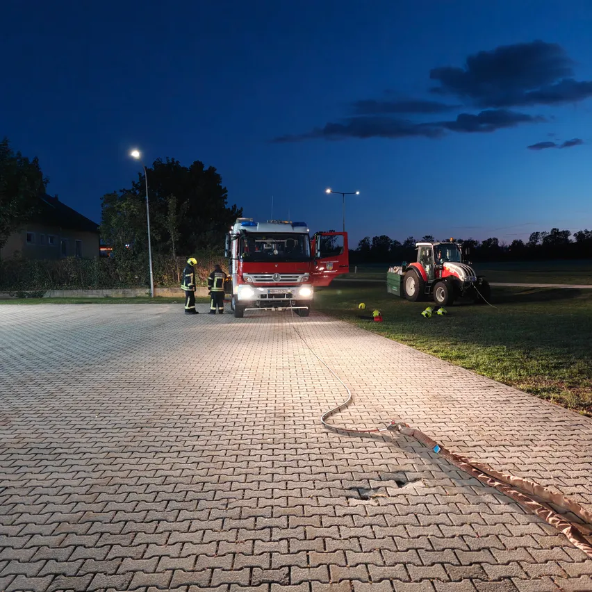 Zwei Feuerwehrleute stehen neben einem Feuerwehrauto mit offener Tür auf einer gepflasterten Straße bei Nacht. Ein Traktor ist in der Nähe geparkt.