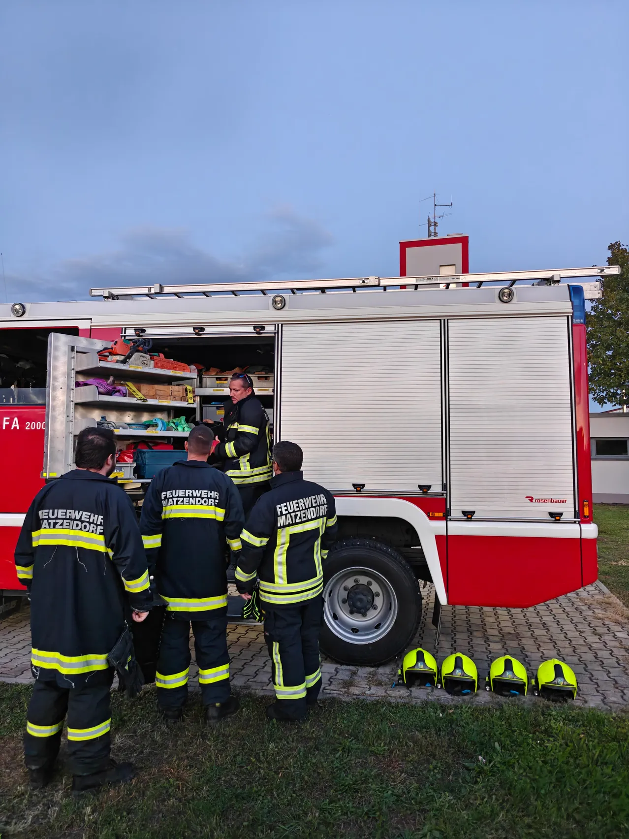 Vier Feuerwehrleute in Uniform stehen um einen Feuerwehrwagen, einer überprüft im Inneren Ausrüstung. Der Wagen steht auf einem Ziegelweg, und Helme sind in der Nähe abgestellt.