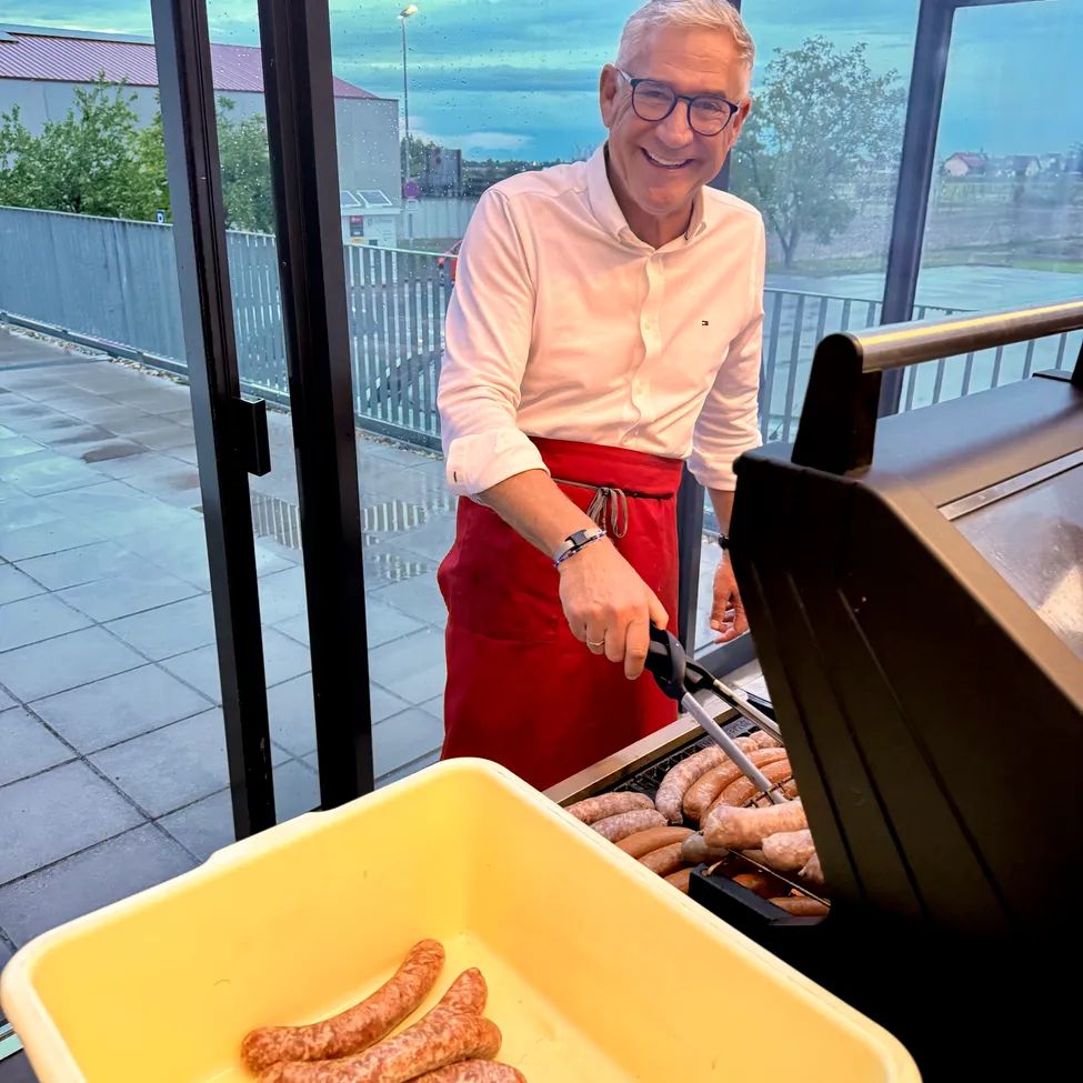 Ein Mann in einer roten Schürze grillt Würste auf einem Grill auf einem Balkon. Er lächelt und hält eine Zange.