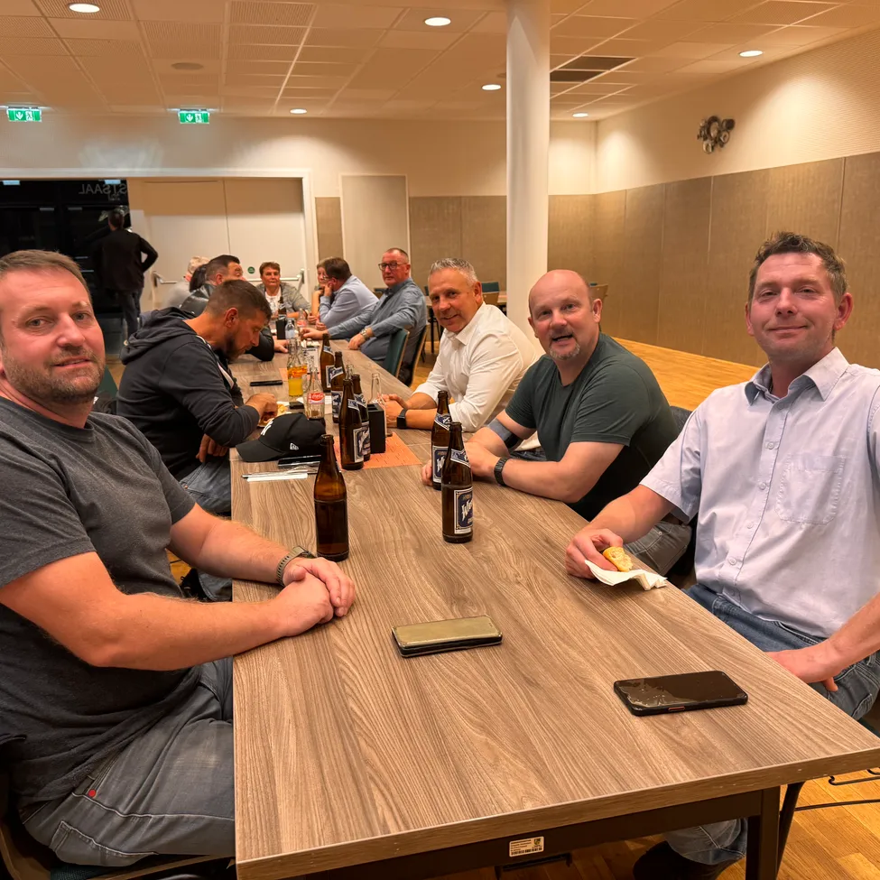 Eine Gruppe von Männern sitzt an einem langen Tisch in einem Raum, lächelt und posiert für ein Foto. Flaschen und Essen sind auf dem Tisch.