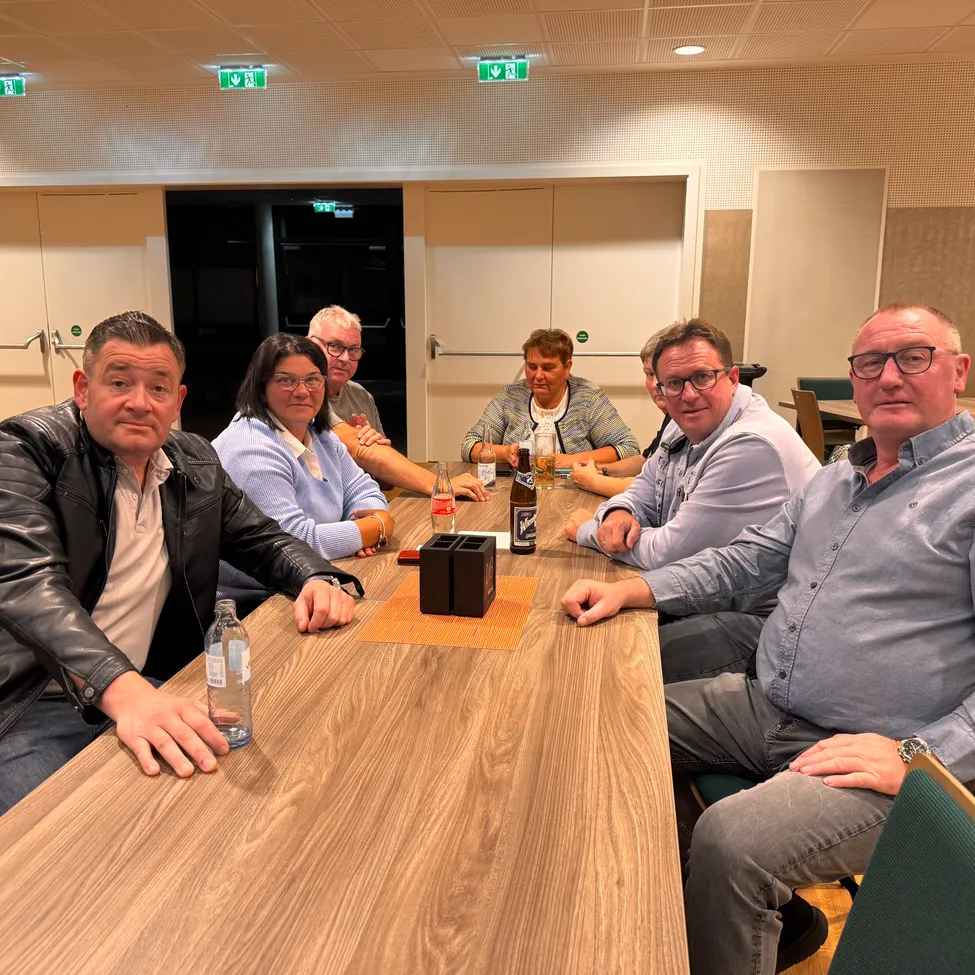Eine Gruppe von Menschen sitzt in einem Besprechungsraum um einen Tisch, einige tragen Brillen und Lederjacken. Flaschen und ein Glas stehen auf dem Tisch.