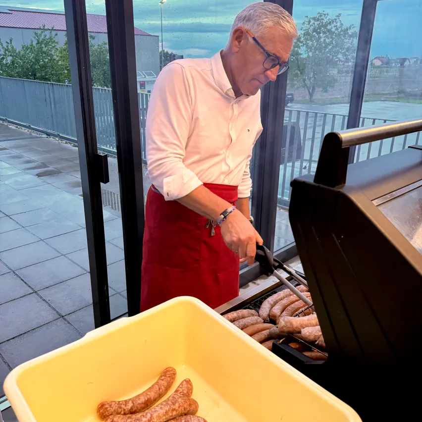 Ein älterer Mann in einem weißen Hemd und einer roten Schürze grillt Würste auf einem Grill im Freien.