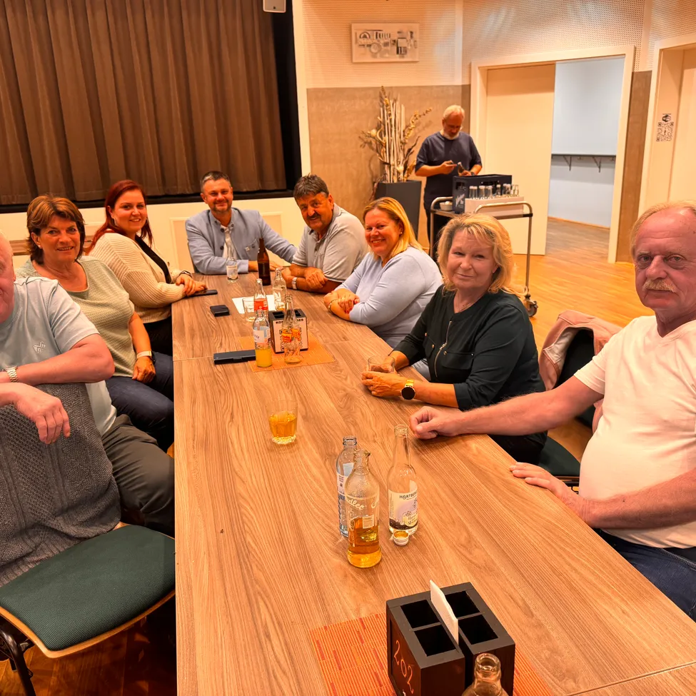 Eine Gruppe von Menschen sitzt um einen Holztisch herum, lächelt und posiert für ein Foto. Flaschen und Gläser stehen auf dem Tisch. Ein Mann steht neben einem Wagen mit Flaschen.