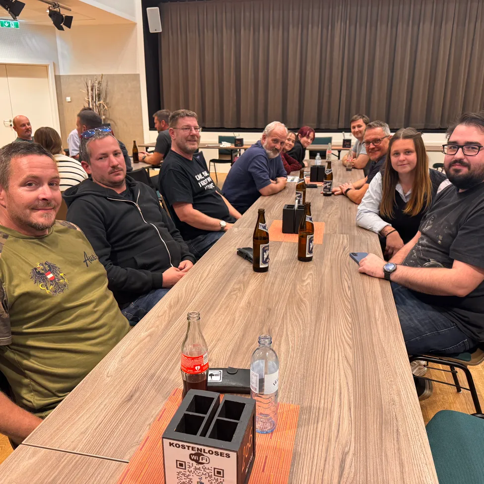 Eine Gruppe von Menschen sitzt um einen Tisch in einem Raum, mit Flaschen und einer Box auf dem Tisch. Sie lächeln und scheinen für ein Foto zu posieren.