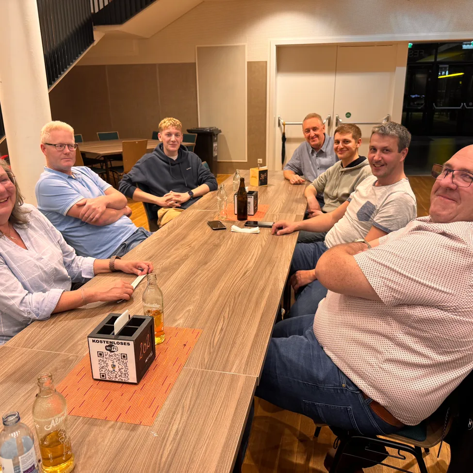 Eine Gruppe von Menschen sitzt um einen Tisch in einem Raum, lächelt und posiert für ein Foto. Flaschen und eine Schachtel sind auf dem Tisch.