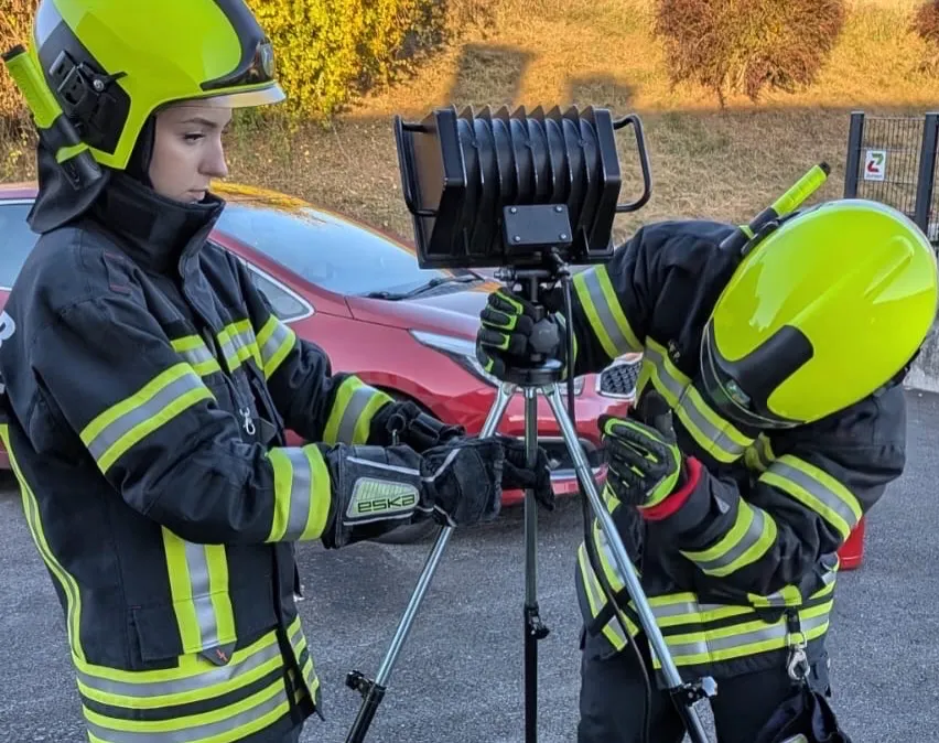 Zwei Feuerwehrleute in voller Montur richten eine Kamera auf einem Stativ in einem Parkplatz ein.