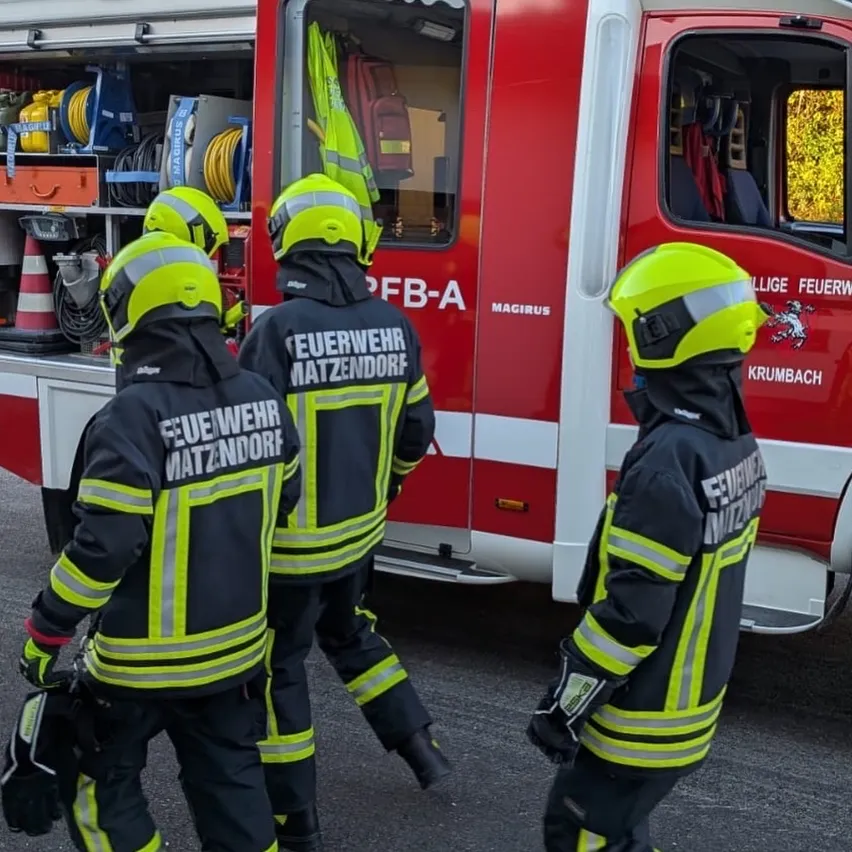 Drei Feuerwehrleute in gelben und schwarzen Uniformen stehen vor einem roten Feuerwehrauto mit dem Wort 'Feuerwehr' darauf.