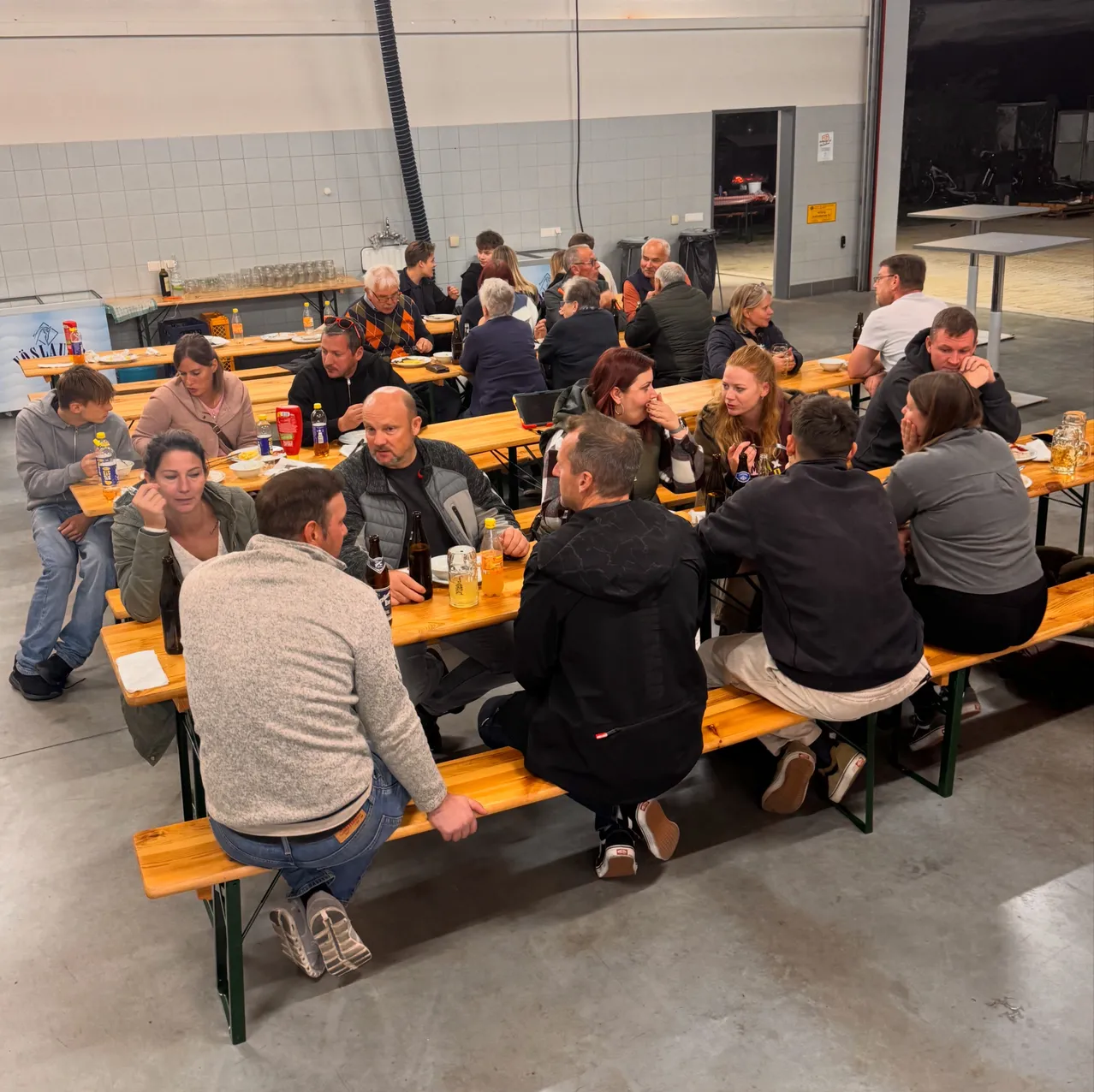 Eine Gruppe von Menschen sitzt an einem langen Holztisch in einem Raum, möglicherweise beim Essen. Einige von ihnen halten Gläser und Flaschen.