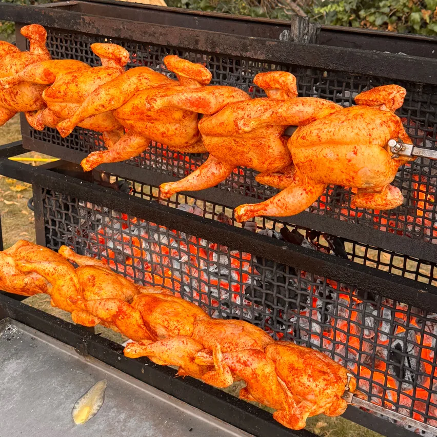 Ein Nahaufnahme eines Grills mit mehreren Hähnchen, die darauf gekocht werden. Die Hähnchen sind goldbraun und haben eine knusprige Haut.