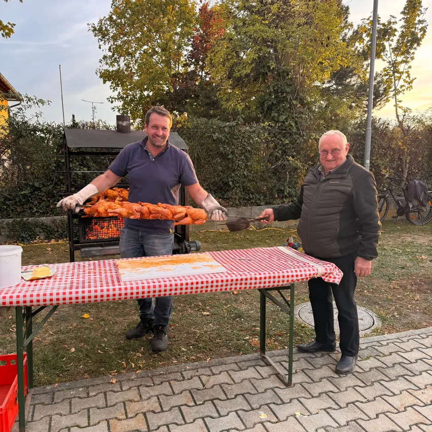 Zwei Männer stehen hinter einem Tisch mit einer rot-weiß karierten Tischdecke. Ein Mann hält ein langes Stück Essen, möglicherweise ein Hähnchenbein, und der andere Mann hält eine Schöpfkelle. Sie lächeln und scheinen für ein Foto zu posieren. Hinter ihnen steht ein Grill mit Feuer und einem Fahrrad.