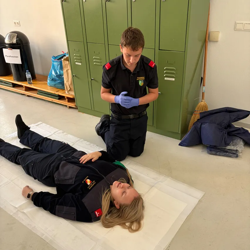 Ein junger Mann in Uniform kniet auf dem Boden neben einer Frau, die auf einem weißen Laken liegt. Die Frau trägt eine Uniform und hat blonde Haare. Hinter ihnen stehen grüne Schließfächer und ein Mülleimer.