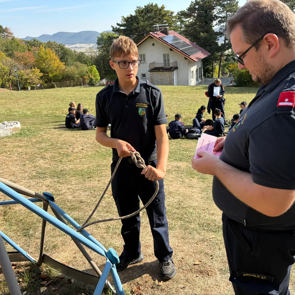 Ein junger Mann in Uniform demonstriert einem Mann in einem blauen Hemd einen Knoten. Sie stehen auf einem Feld mit einem Haus und Bäumen im Hintergrund.