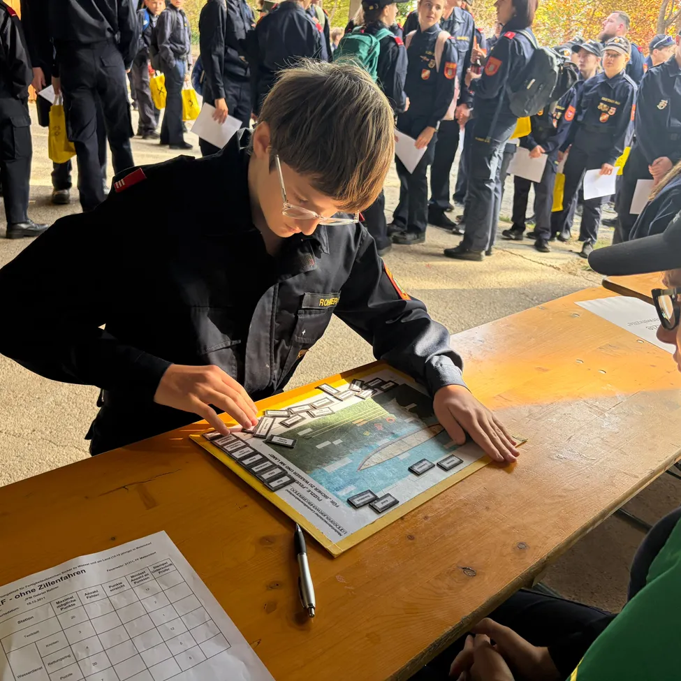 Ein Junge in Uniform arbeitet an einem Brettspiel mit einer Karte darauf. Er sitzt an einem Tisch mit einem Stift und einem Papier mit einem Formular. Eine andere Person sitzt neben ihm. Hinter ihnen stehen Leute in Uniform in einer Reihe.