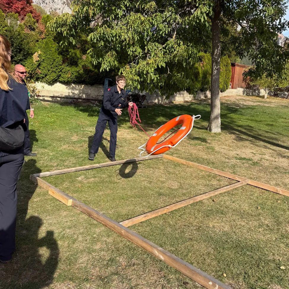 Eine Person in Uniform zieht an einem Seil, an dem ein orangefarbener Rettungsring an einem Holzrahmen auf einem Grasfeld befestigt ist.