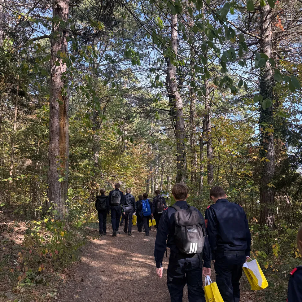 Eine Gruppe von Menschen geht auf einem Pfad im Wald, trägt Rucksäcke und gelbe Taschen.