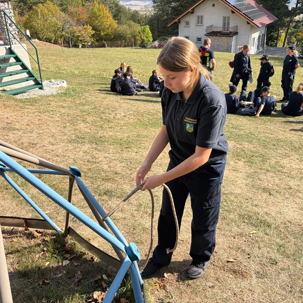 Eine Frau in einer blauen Uniform repariert eine blaue Struktur auf einem Feld. Hinter ihr sitzen mehrere Personen auf dem Gras, möglicherweise in einer Besprechung.