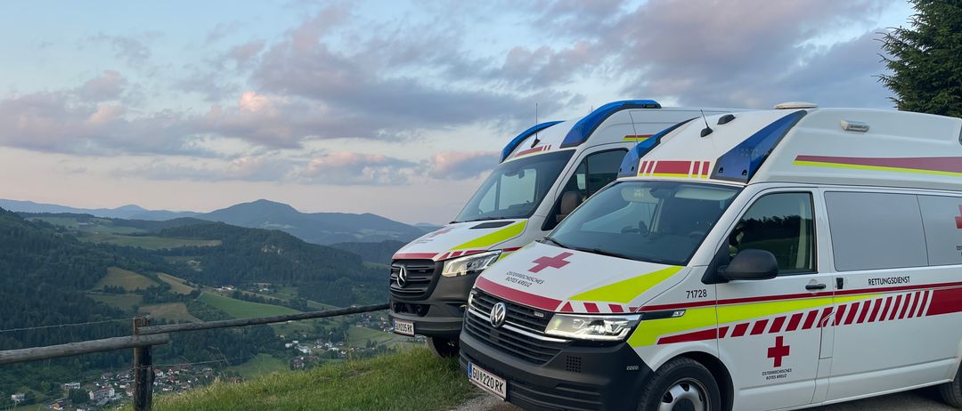 Zwei Krankenwagen sind auf einer Straße geparkt, einer hinter dem anderen. Sie haben rote und weiße Streifen und rote Kreuze an den Türen. Die Straße ist von einem Metallgeländer umgeben. Dahinter befindet sich ein grasbewachsener Hügel mit Blick auf Berge. Der Himmel ist blau mit einigen Wolken.