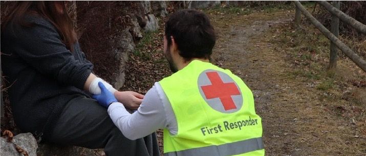 A first responder is attending to a person. They are wearing a reflective vest with a red cross. The event is on April 18, 2026, from 6:00 PM to 7:00 PM.