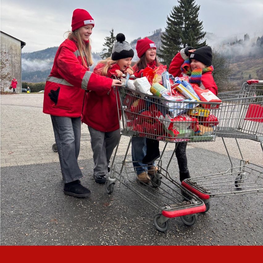 Eine Gruppe von Menschen, die rote Jacken tragen, schiebt einen mit Gegenständen gefüllten Einkaufswagen, möglicherweise für eine Spende. Sie stehen auf einem gepflasterten Bereich in der Nähe eines Gebäudes.