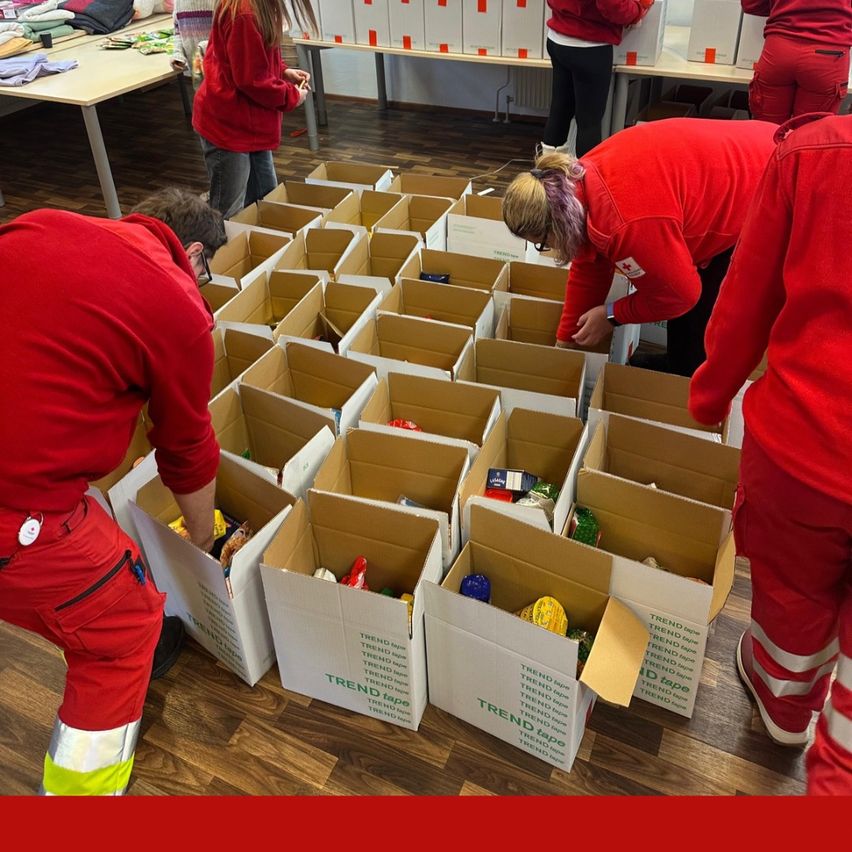 Freiwillige in roten Uniformen sortieren Gegenstände in Kartons mit der Aufschrift TREND tape. Eine Frau mit Brille beugt sich über einen Karton. Sie befinden sich in einem Raum mit Holzböden.
