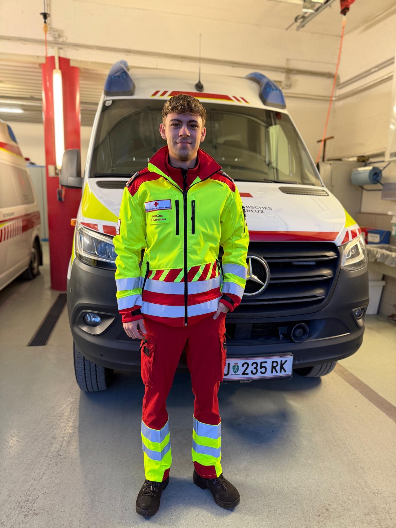 Ein junger Mann in einer reflektierenden Sicherheitsjacke steht vor einem Krankenwagen in einer Garage.