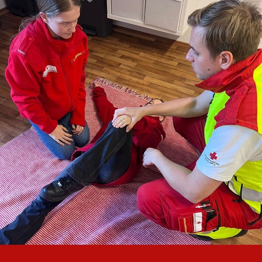 Eine Frau in einer roten Uniform kniet neben einem Mann in Rot und Schwarz und hält dessen Bein fest. Der Mann trägt eine rot-gelbe Sicherheitsweste. Beide sind drinnen auf einem rot-weißen Teppich.