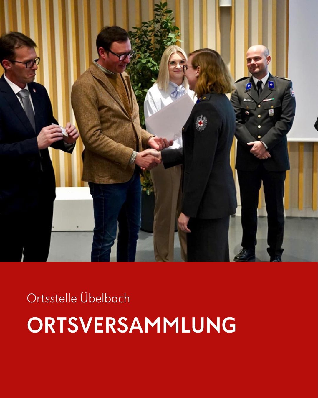 Eine formelle Veranstaltung mit fünf Personen in Anzügen und Uniformen, die Hände schütteln. Ein Mann in Uniform übergibt einem Frau ein Papier. Dahinter stehen zwei Männer in Anzügen, einer hält ein Papier. Der Hintergrund ist gelb gestreift mit einer Pflanze.