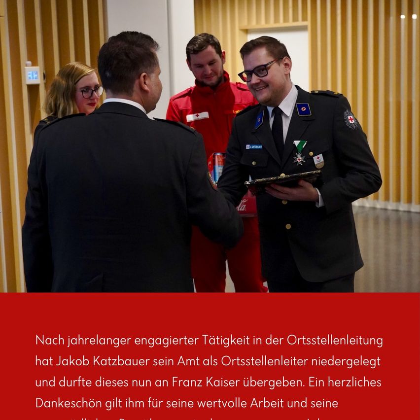 Eine formelle Zeremonie zeigt einen Mann in Uniform, der einem anderen Mann in Anzug eine Plakette überreicht. Dahinter stehen eine Frau und ein weiterer Mann in rotem Uniform. Die Umgebung ist indoor mit Holzwänden und einem roten Hintergrund.