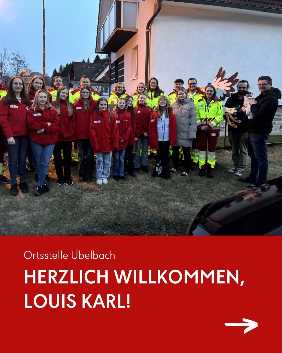 Eine Gruppe von Menschen in roten und gelben Jacken steht vor einem Haus und begrüßt ein Neugeborenes. Die Gruppe lächelt und posiert für ein Foto.