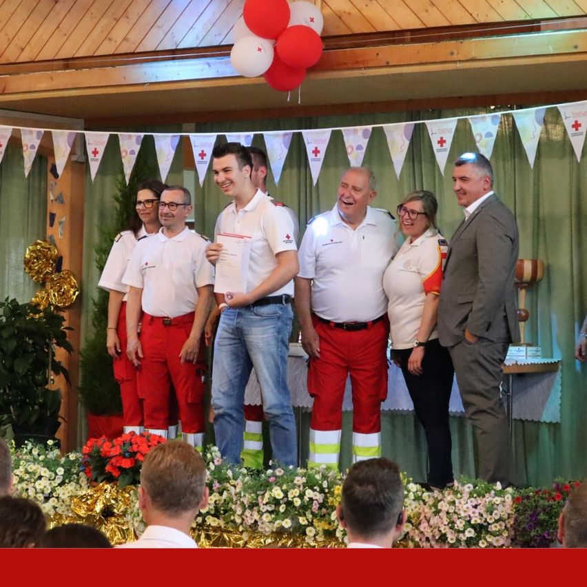 Eine Gruppe von Menschen in roten und weißen Uniformen steht auf einer Bühne mit Luftballons und Blumen vor ihnen. Sie scheinen für ein Foto zu posieren.