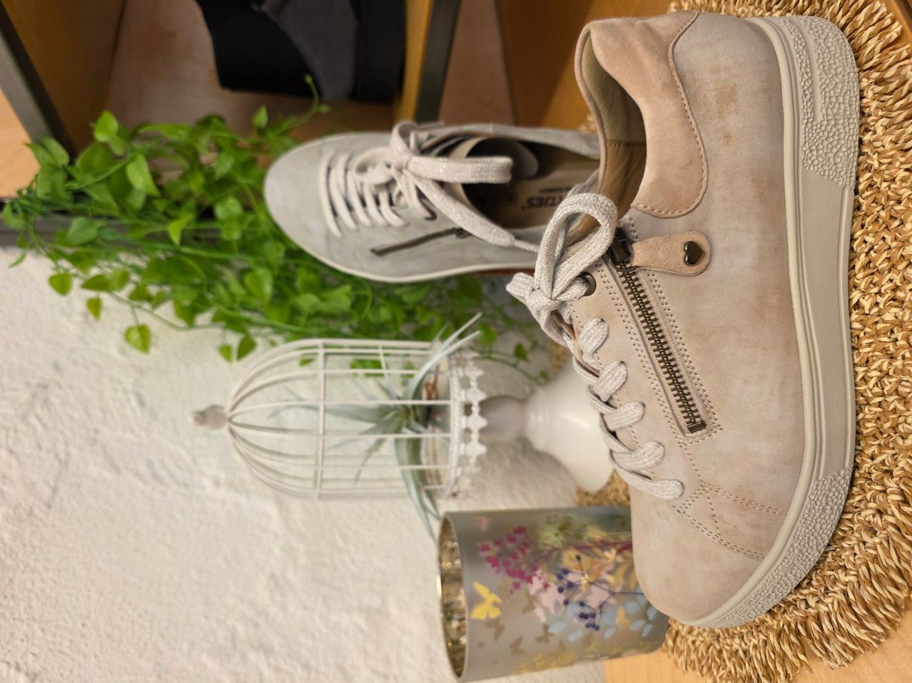 Ein Paar beige Sneakers mit einem herzförmigen Reißverschlussdetail. Auf einem weißen Tuch platziert, neben einem weißen Vogelkäfig und einem dekorativen Gegenstand.
