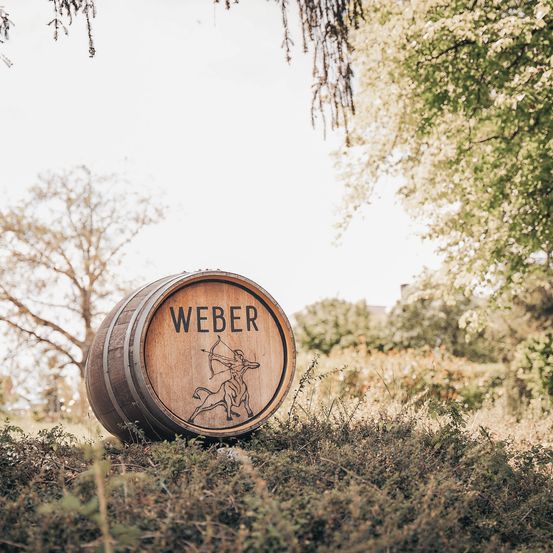 Ein Holzfass mit dem Wort Weber und einer Illustration eines Zentaurs mit Bogen und Pfeil steht draußen zwischen Büschen und Bäumen.