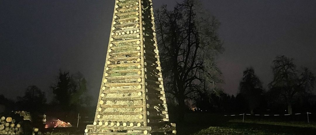 Ein hoher Holzstapel mit einem Weihnachtsbaum auf der Spitze ist bei Nacht in einem offenen Feld beleuchtet.