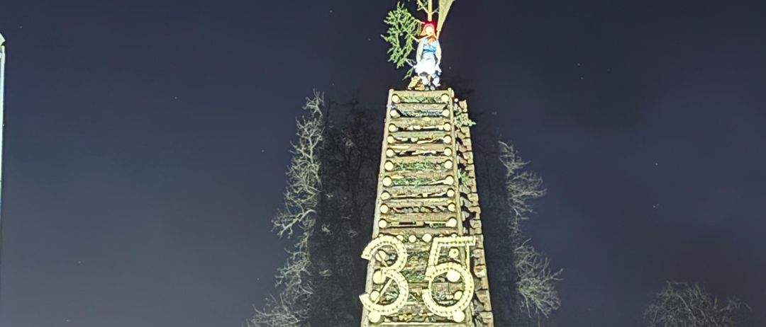 Eine nächtliche Szene zeigt eine hohe Struktur mit '35' darauf, gekrönt von einem Baum und einer Figur, beleuchtet vor einem dunklen Himmel mit entfernten Bäumen.