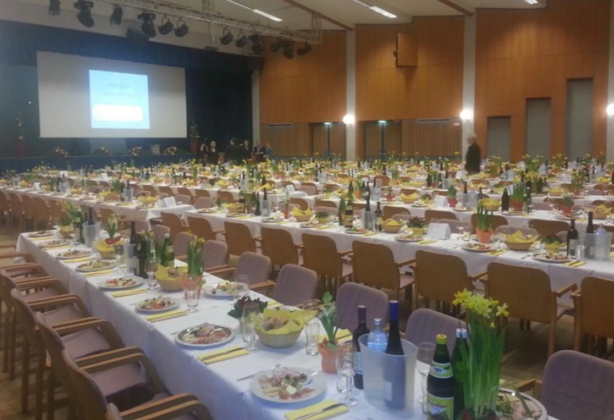 Ein großer Saal mit vielen für ein Bankett gedeckten Tischen. Jeder Tisch hat Teller, Weinflaschen und Blumen. Menschen stehen in der Nähe der Bühne, und eine Projektionsleinwand ist sichtbar.
