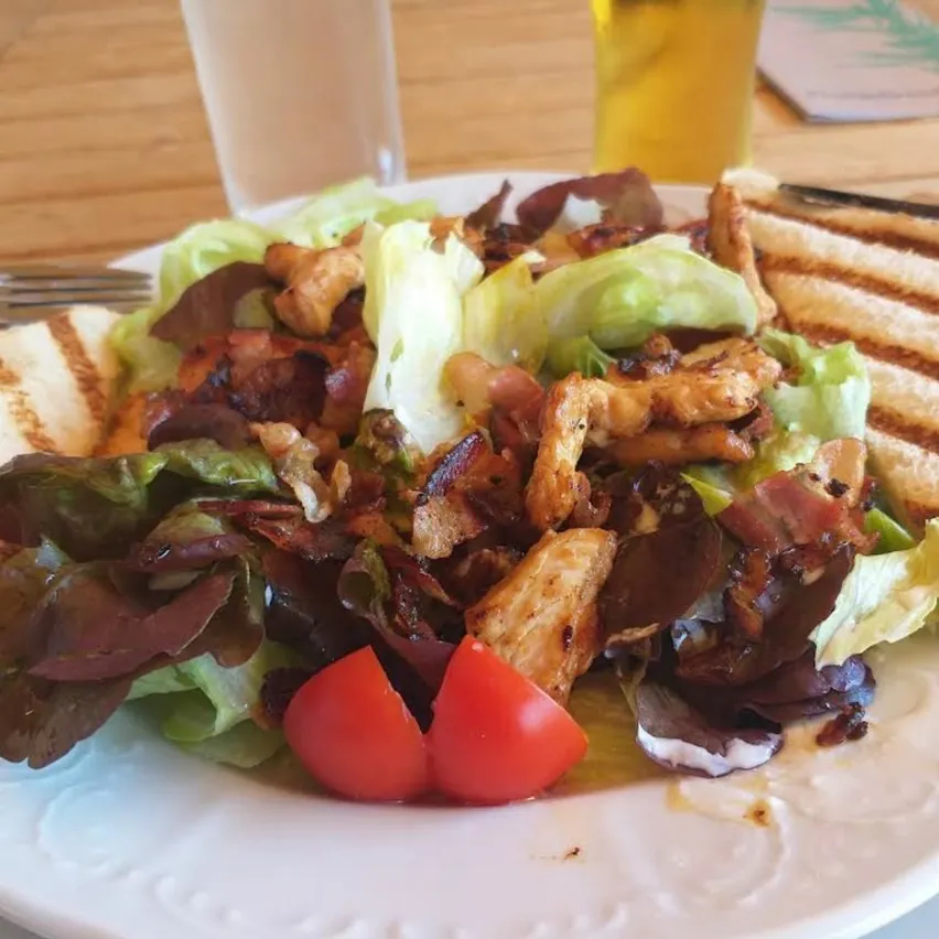 Ein weißer Teller mit einem Salat, bestehend aus Salat, Tomaten und gegrilltem Hähnchen, begleitet von einem Glas Bier und einem Glas Wasser auf einem Holztisch.