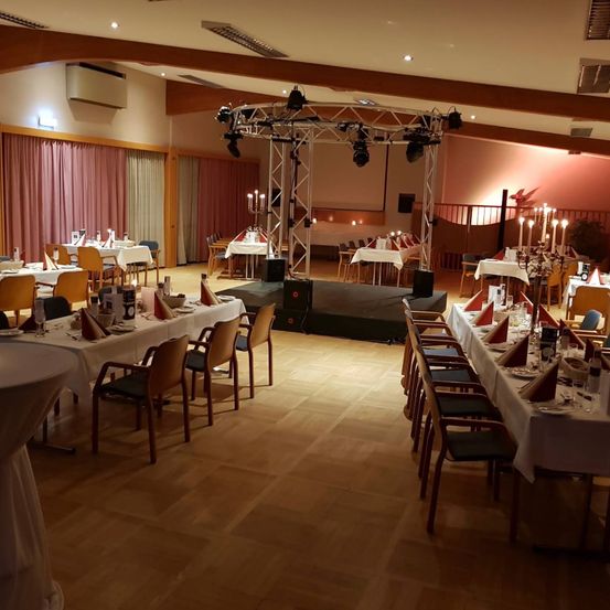 Ein leerer Bankettsaal mit Holzboden. Es gibt eine Bühne mit einem Metallrahmen, einem Kerzenständer und einem Tisch, der für eine Mahlzeit gedeckt ist. Es gibt Stühle um die Tische herum angeordnet.