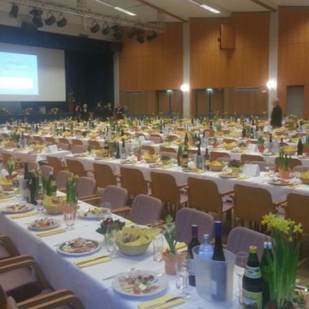 Ein Bankettsaal ist eingerichtet mit langen Tischen, die mit weißen Tischdecken bedeckt sind und Teller mit Essen, Weinflaschen und gelben Blumen halten. Menschen stehen in der Nähe einer Projektionsleinwand vorne.