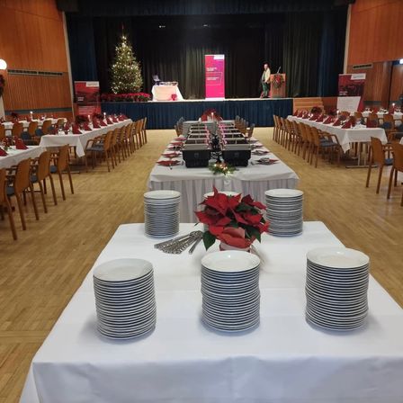 Ein Raum für ein Bankett mit Tischen und Stühlen, geschmückt mit Weihnachtsdekoration. Teller, Besteck und ein Blumen-Centerpiece sind auf den Tischen arrangiert. Eine Bühne im Hintergrund zeigt einen Weihnachtsbaum und ein festliches Banner.