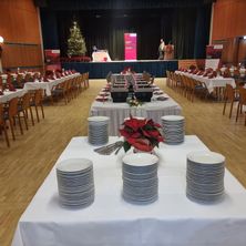 Ein Raum für ein Bankett mit Tischen und Stühlen, geschmückt mit Weihnachtsdekoration. Teller, Besteck und ein Blumen-Centerpiece sind auf den Tischen arrangiert. Eine Bühne im Hintergrund zeigt einen Weihnachtsbaum und ein festliches Banner.