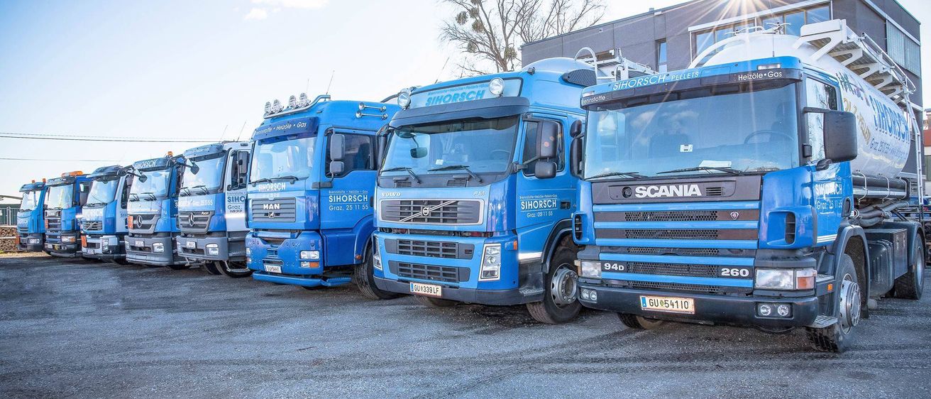Eine Reihe blauer Lastwagen mit dem Schriftzug Sihorsch davor steht vor einem Gebäude unter einem klaren Himmel.