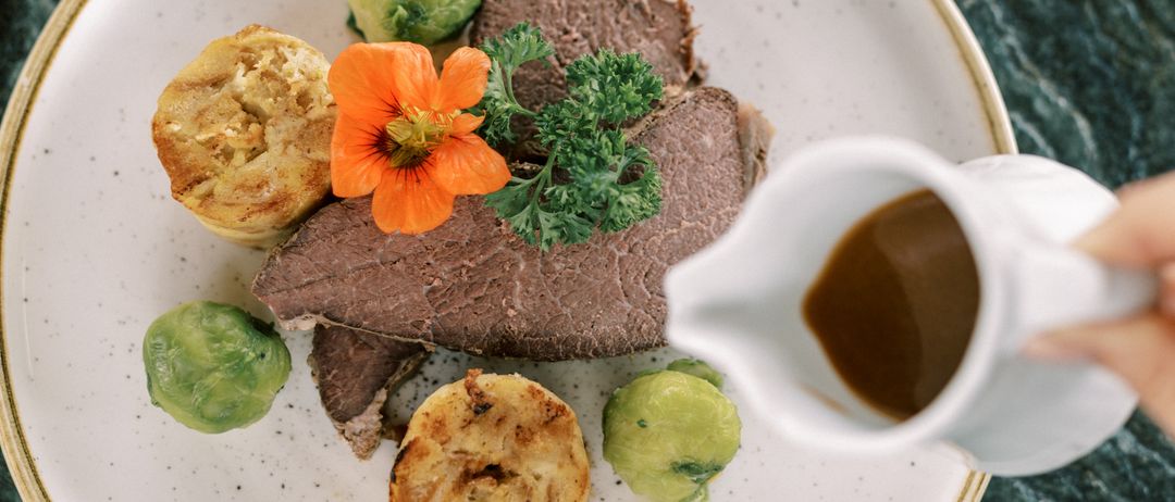 Eine Hand gießt Soße über einen Teller mit Roastbeef, Yorkshire Pudding, Rosenkohl und einer orangefarbenen Blume, mit einem Glas Wein in der Nähe.