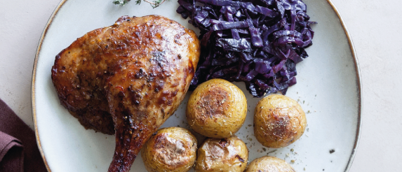 Ein Teller mit einem gebratenen Hähnchenschenkel, gebratenen Kartoffeln und rotkohl auf einer weißen Oberfläche.