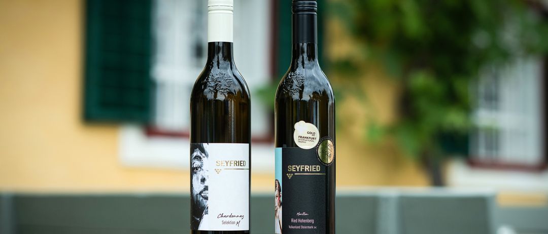 Zwei Seyfried-Weinflaschen, eine weiße und eine rote, stehen auf einem Fass vor einem Haus.