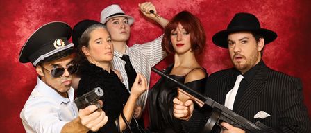 Fünf Personen in Vintage-Gangster-Kleidung posieren vor einem roten Hintergrund. Ein Mann und eine Frau halten Pistolen, während ein anderer Mann einen Stock hält. Die anderen schauen mit verschiedenen Ausdrücken zu.