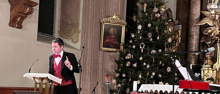 Ein Mann steht in einer Kirche an einem Podium, trägt eine rote Fliege und Weste, mit einem Weihnachtsbaum, Musikinstrumenten und einer Statue dahinter.