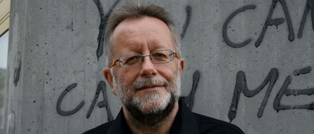 Ein älterer Mann mit grauem Haar und Bart steht vor einer mit Graffiti bedeckten Wand, trägt eine Brille und ein schwarzes Hemd.