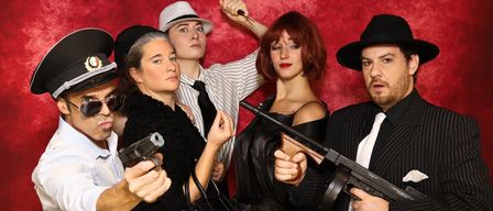 Five people dressed in 1920s gangster attire pose with guns for a photograph against a red background.