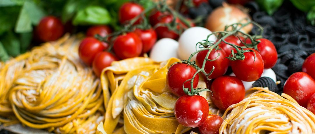 Ein Nahaufnahme von verschiedenen frischen Zutaten, darunter gelber Pasta, rote Tomaten, grüne Blätter und weiße Eier. Die Zutaten sind ordentlich auf einer Holzoberfläche angeordnet.